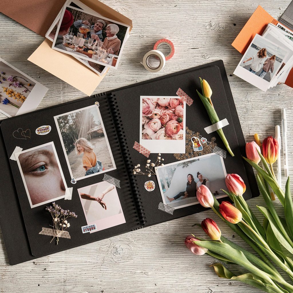 Image of an open, black-colored photo album with taped photos inside and various flowers, doodles, and stickers scattered about. The photos inside consist of a smiling blonde woman, a close-up of a blue eye, two women sitting and laughing, a close-up of pink roses, and two hands holding each other.
